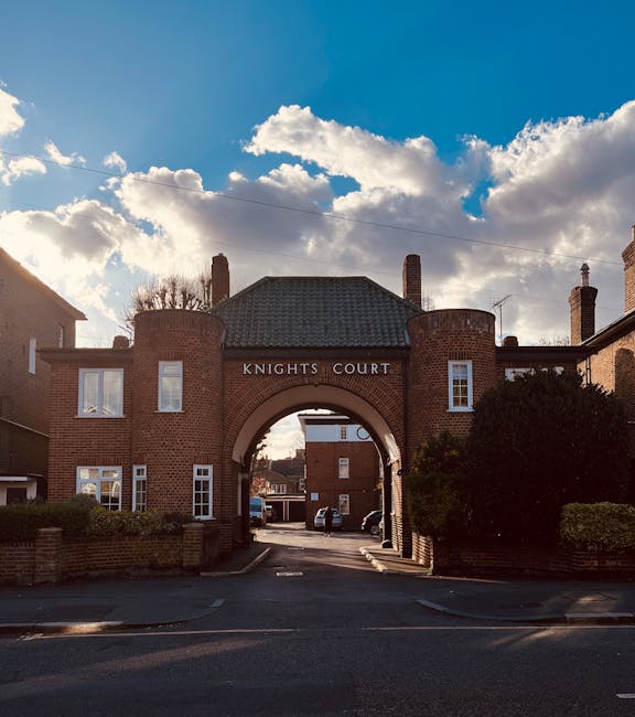 A brick residential building with a large central archway labeled 'Knights Court' provides access to a narrow street or courtyard, visible under a partly cloudy sky with sunlight casting shadows on the pavement. To the left and right of the archway, there are windows with white frames and small bushes along the perimeter wall. Inside the arch, parked cars can be seen, indicating a private parking area or service zone. The image captures a typical urban environment suitable for house removals and furniture transport, highlighting a location where narrow Kingston streets may present access challenges for larger vehicles. This setting is relevant for relocation services provided by companies like Man and a Van Kingston, which handle loading and unloading processes during home removals or furniture transport, especially in areas with restricted street access. The scene emphasizes the importance of planning for narrow street mobility in local property relocations.