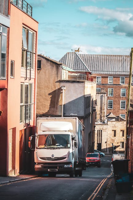 A white delivery van parked on a narrow Kingston street with a loading area at the rear, surrounded by residential buildings of varying heights and architectural styles, including a modern peach-colored apartment on the left and older brick structures in the background. The street is paved with asphalt and features double yellow lines along the edge, with a small black wheelie bin on the right sidewalk. The van is positioned close to the curb, preparing for home relocation or furniture transport, while a car is visible further down the street. The sky is partly cloudy, and the lighting suggests late morning or early afternoon. The scene illustrates typical urban moving logistics near Kingston's narrow streets, which may pose access challenges for larger vehicles, a situation that Man and a Van Kingston often manages through strategic loading and unloading processes during house removals or packing and moving services.