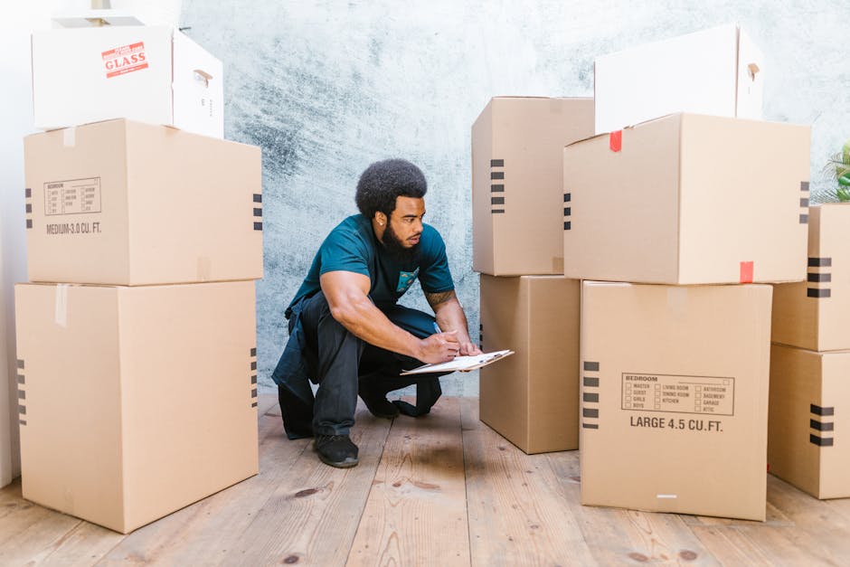 A man from Man and a Van Kingston, dressed in a dark blue t-shirt and black trousers, is kneeling on a wooden floor inside a room, surrounded by large cardboard boxes used for home relocation. He is holding a clipboard and appears to be inspecting or making notes on a packing checklist. The boxes are labelled with size and contents, with some sealed with packing tape, and are stacked neatly around him, indicating an organized moving process. The room has a textured grey wall and is well-lit, with natural light coming from behind the camera. In the background, additional boxes and a glimpse of an entryway or doorway suggest the ongoing packing and furniture transport involved in a house move using professional services like those offered by Man and a Van Kingston.