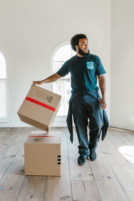 A man with an afro hairstyle and a beard is inside a bright, minimally decorated room with white walls and wooden flooring. He is wearing a navy blue T-shirt with a logo on the chest and dark trousers, and he is in the process of lifting or placing a cardboard box with red tape across the top. The man is standing near two cardboard boxes, one positioned on the floor and the other being held in one hand, suggesting a home relocation or furniture transport activity as part of a packing and moving process. Behind him, a large arched window allows natural light to fill the space, highlighting the clean, uncluttered environment. This scene illustrates the logistics involved in house removals, with the man collaborating in packing or loading items for a move. The focus on boxes, the interior setting, and the careful handling of packaging materials reflect professional removals and moving services handled by companies like Man and a Van Kingston.
