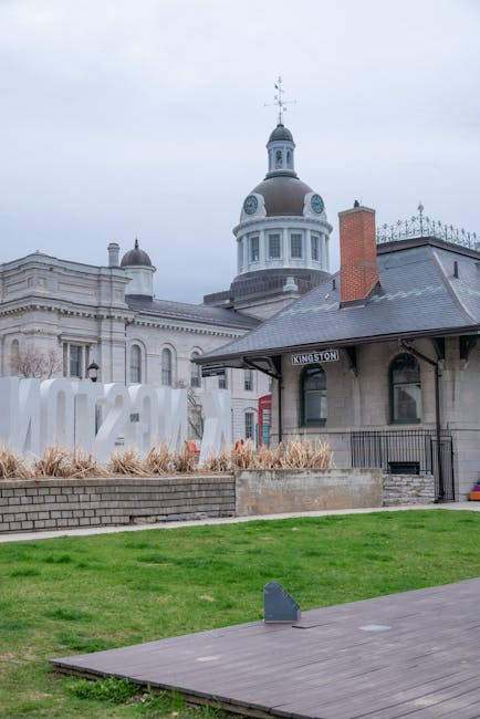 The image shows the exterior of a historic building with a prominent dome and ornate architectural details, situated behind a landscaped area with grass, dry ornamental plants, and a low brick wall. In the foreground, a wooden pathway leads past a small, rectangular grey object on the pavement. To the right, part of a small, single-story building with a sign reading 'KINGSTON' is visible, featuring dark window frames and a sloped roof. The sky is overcast, providing diffused natural light. In the setting where house removals or furniture transport services might take place, the scene captures a typical urban location likely near a train station or town centre, with visible adjacent historic structures. This context aligns with loading or moving activities in a city environment, where Man and a Van Kingston could facilitate home relocations or furniture deliveries, exemplified by the professional scene of an urban move preparing for transport.