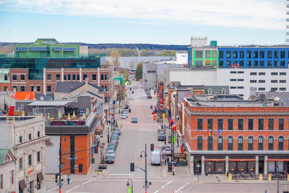 An urban street scene with a view looking down a busy city street towards the river in the background, featuring a variety of commercial buildings, both modern and historic, with storefronts, signage, and windows. On the street, parked cars line both sides, and pedestrians are visible walking along the sidewalks. In the foreground, there are several trucks and vans, including a large white van, associated with furniture transport and home relocation services, with some loading or unloading boxes and furniture protected by plastic wrap and blankets. The vehicles are positioned near the entrances of buildings, suggesting active parts of a moving or delivery process. The environment is well-lit with natural daylight, and the scene captures the logistical elements involved in house removals within a city setting. Occasionally, signage and street furniture support the urban context, aligning with professional removals and transportation services such as those offered by Man and a Van Kingston.