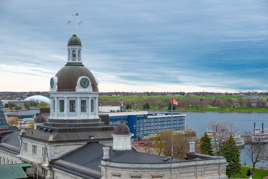 The image shows an urban waterfront scene with a historic building featuring a large clock tower topped by a domed roof and weather vane. The building's architecture includes white stonework, arched windows, and decorative features, situated on a paved area near the water. In the foreground, the edge of a rooftop with dark grey roofing and chimneys is visible. The background displays a large river or lake, surrounded by green parks and trees, with several boats docked along the shoreline. Across the water, modern blue buildings and additional greenery can be seen, along with an overcast sky filled with layered grey-blue clouds. The scene captures a mix of historical and contemporary structures, set against a natural landscape, reflecting elements relevant to home relocation and the logistics involved in furniture transport. The presence of boats and urban development emphasizes the movement and shipping aspects associated with moving services by [COMPANY_NAME], aiding in a professional house removal or furniture transport process from Kingston to other locations.