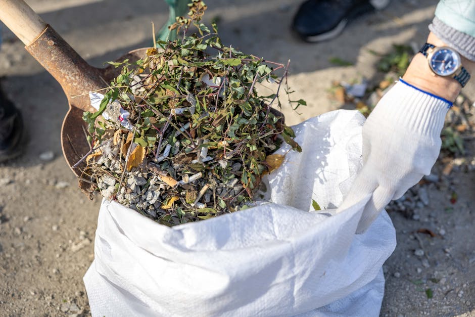A person wearing a white glove is using a shovel to load a white sack with garden waste, including small branches, leaves, and gravel, during a home relocation or outdoor clearance process. The sack is positioned on the ground outdoors, with gravel and dirt visible. The individual’s wristwatch and sleeve are partially visible, indicating a casual outfit. This scene reflects the packing and loading activities involved in removals services, such as clearing garden debris before transportation. The setting suggests a residential or property exterior environment, where items are being prepared for removal in compliance with local waste rules, as addressed on the Kingston council bulky waste rules page. Man and a Van Kingston provides professional removals services that include efficient loading and transportation of various household and garden items as part of their house move or clearance operations.