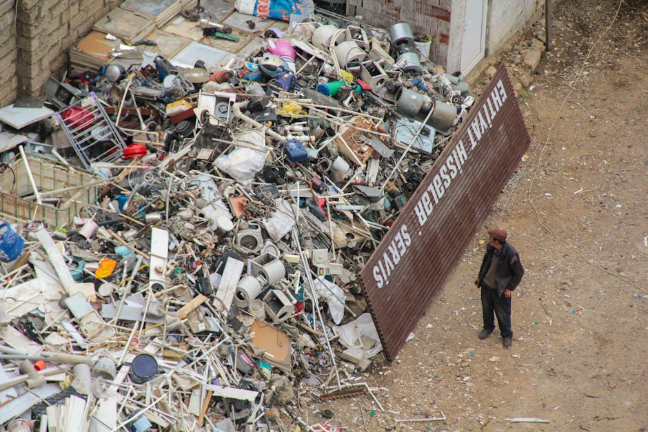A man dressed in dark clothing and a brown hat stands outdoors on a patch of bare earth near a large pile of assorted household waste and discarded items, including broken appliances, plastic bottles, metal frames, cardboard boxes, and miscellaneous debris. Adjacent to the waste pile is a large brown metal sign bearing white text that reads 'FINAL WASTE SERVICES,' which is placed at an angle. The waste appears to be contained in a designated area for disposal or recycling, with some items spilling onto the surrounding ground. The setting is an open outdoor space, likely near a building or waste management facility, with the waste collection process visible as part of the household clearance or home relocation activity. This scene is associated with the manpower and logistics involved in furniture transport, packing, and moving services, as managed by Man and a Van Kingston, in compliance with local waste removal regulations.