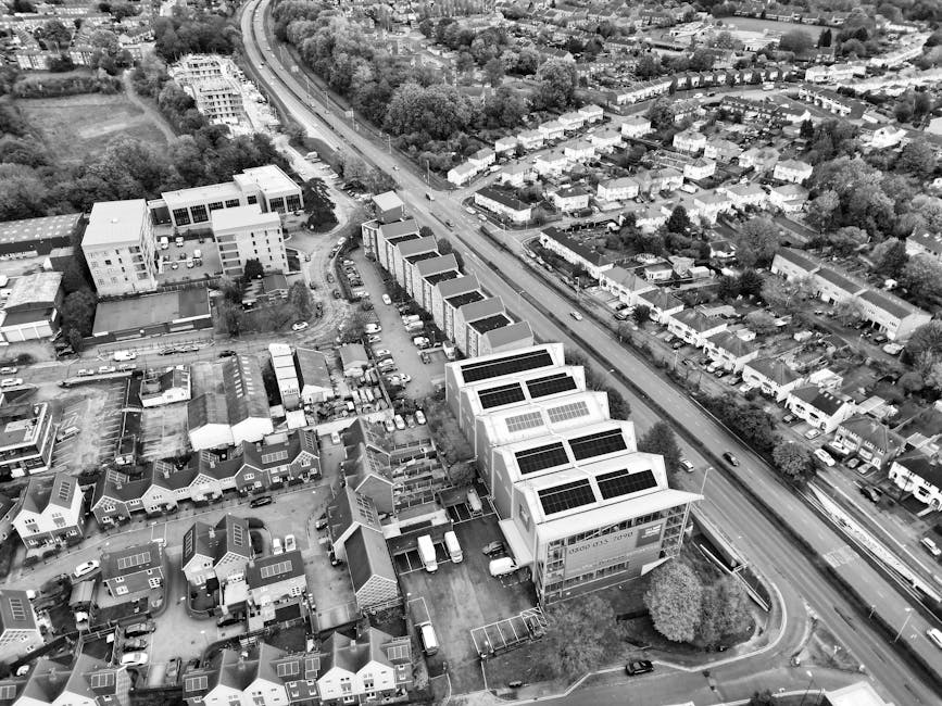 An aerial black-and-white photograph shows a line of modern, cube-shaped buildings with flat roofs fitted with solar panels, situated along a paved area near a busy road with moving vehicles. Adjacent to the buildings, there is a parking area filled with several cars. Surrounding the property are smaller residential houses, some with solar panels on their roofs, and green spaces with trees. The scene appears to depict a home relocation process or furniture transport activity associated with a house removal service, possibly involving the packing and loading of belongings. The image provides a clear view of the urban environment and the logistical layout for moving services, which could be coordinated by Man and a Van Kingston as part of their removals offering for student moves, such as those related to Kingston University Penrhyn Road.