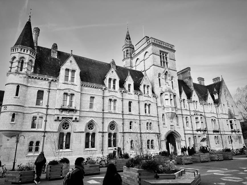 Black and white photograph of a large historic stone building with multiple towers, turrets, and gabled roofs, characteristic of castle or gothic architecture, situated on a university campus. The front façade features arched windows, some with decorative tracery, and an entrance with a rounded arch doorway. The exterior walls are textured stone with detailed masonry work. In the foreground, there are several large rectangular wooden planters filled with plants, and people are walking along the paved area in front of the building, with bicycles parked nearby. The scene appears to be daytime with natural light illuminating the structure and surroundings. This image relates to house and property removals, possibly indicating a move involving the historic building or a university-related relocation service, as handled by companies like Man and a Van Kingston, specialising in home relocation and furniture transport.