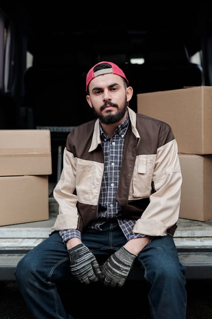 A young man with a beard, wearing a red baseball cap backwards, a beige and brown jacket over a checked shirt, sitting on the lower edge of an open van’s cargo area during a home relocation. Surrounding him are several large cardboard boxes, some partially wrapped in plastic or packing materials, ready for transportation. The background shows the dark interior of the van, indicating it is part of a furniture transport or packing and moving process as part of house removals by Man and a Van Kingston. The scene captures a moment of preparation in the loading process, with the man appearing calm and attentive, indicating careful handling of household items for a move to Canbury Gardens, Kingston.