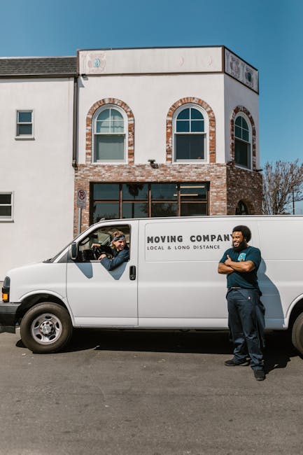 A white moving van with the branding 'MOVING COMPANY, LOCAL & LONG DISTANCE' parked on a street in front of a two-story residential building. One man with light hair, wearing a beanie and dark clothing, sits in the driver's seat and appears to be in conversation with a second man standing outside the sliding door of the van. The second man has dark skin, curly hair, and is dressed in a dark shirt and trousers, with arms crossed, smiling attentively. The scene suggests active loading or unloading as part of a house removal or relocation process, with the van positioned close to the property for efficient furniture transport and packing during a home move. The building in the background features a brick façade with large arched and rectangular windows, and the weather is clear with bright sunlight. Man and a Van Kingston, as a professional removals service, is evident in the context of packing, loading, and transporting household belongings effectively.