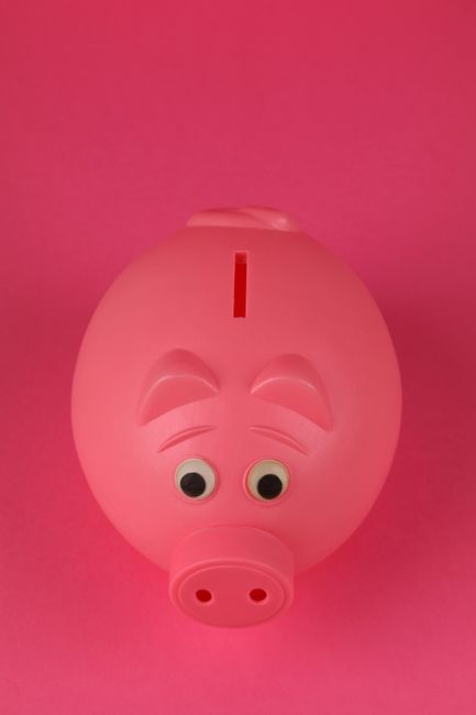 A photograph featuring a person's hand inserting a pound coin into a black ceramic piggy bank positioned on a white surface covered with various loose coins, including copper, silver, and gold-colored
