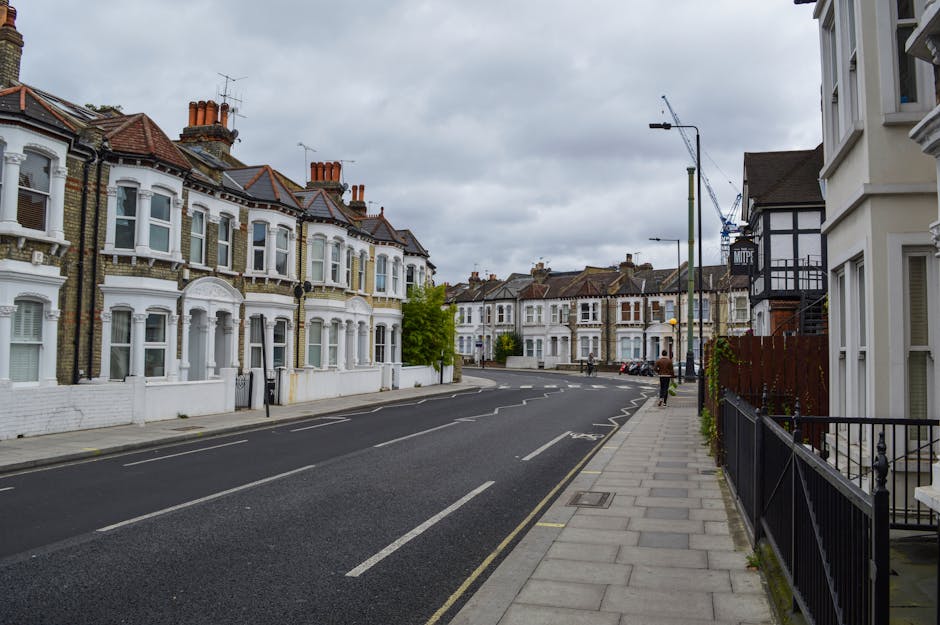 A quiet street in Kingston KT1 with a row of Victorian-style terraced houses featuring bay windows, brickwork, and terracotta chimney pots on the roofs. The pavement runs alongside the houses, with a black metal fence bordering one property and a small amount of greenery visible. Overhead, an overcast sky with grey clouds is present. Streetlights and parked cars are seen further down the road, while a construction crane is visible in the background, indicating ongoing development nearby. The scene captures the typical residential environment associated with house removals and local relocation services by Man and a Van Kingston, highlighting the area's architecture and street layout suitable for home relocation activities.