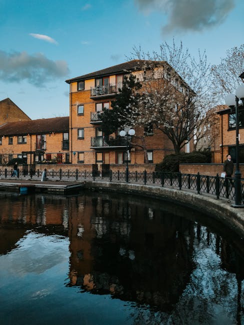 A multi-storey brick residential building situated above a canal or river, with several windows and balconies. The building is surrounded by trees, including one with sparse branches, and a lamp post with multiple spherical lights positioned near the walkway. In the foreground, a paved promenade runs alongside the water, which reflects the building and sky above. A few pedestrians are walking along the path, some of whom appear to be engaged in a house removal or transportation process, possibly related to a home relocation service. Man and a Van Kingston, a company specialising in removals, may be involved in moving furniture or packing materials visible in the scene. The overall setting features natural lighting with partly cloudy skies, enhancing the urban yet calm environment conducive to moving logistics and furniture transport activities.