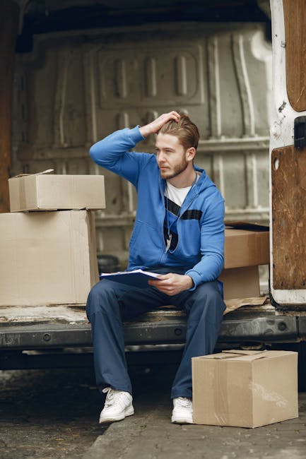 A young man with light skin, wearing a blue hoodie and navy trousers, is seated on the edge of an open van's loading area, surrounded by several cardboard boxes of various sizes. He is holding a clipboard and a pen, with one hand resting on his head, appearing to be taking a break or assessing the packing process during a house removal. The interior of the van shows a metal panel background, indicating a typical moving vehicle used for furniture transport and packing during home relocation. The boxes are placed both inside the van and on the pavement beside him, some sealed with packing tape, suggesting a recent or ongoing packing and loading activity. The scene is outdoors, with natural daylight illuminating the area, and the focus emphasizes the logistics involved in professional removals, as provided by companies like Man and a Van Kingston, specializing in timely and efficient moving services.