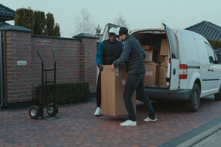 Two men, dressed in casual clothing with padded jackets and caps, are engaged in a home relocation task outside a residential property during daytime. They are carefully lifting a large cardboard box, which is sealed with packing tape, onto or from a white van that is parked on a brick-paved driveway. The van’s rear doors are open, revealing several more cardboard boxes and packing materials stacked inside. An empty two-wheeled hand truck is positioned nearby on the pavement, ready for transportation of additional items. The background features a brick boundary wall, a row of trees, and a residential house with a tiled roof, under a cloudy sky. The scene captures the loading process involved in packing and moving services, likely performed by Man and a Van Kingston, to facilitate house removals or furniture transport. The environment is well-organized, highlighting the logistical aspects of local home relocation and packing procedures.