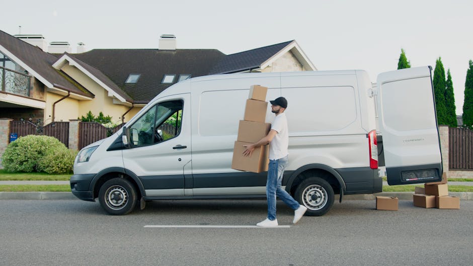 A young man with light skin, wearing a blue hoodie and navy trousers, is seated on the edge of an open van's loading area, surrounded by several cardboard boxes of various sizes. He is holding a clipboard and a pen, with one hand resting on his head, appearing to be taking a break or assessing the packing process during a house removal. The interior of the van shows a metal panel background, indicating a typical moving vehicle used for furniture transport and packing during home relocation. The boxes are placed both inside the van and on the pavement beside him, some sealed with packing tape, suggesting a recent or ongoing packing and loading activity. The scene is outdoors, with natural daylight illuminating the area, and the focus emphasizes the logistics involved in professional removals, as provided by companies like Man and a Van Kingston, specializing in timely and efficient moving services.